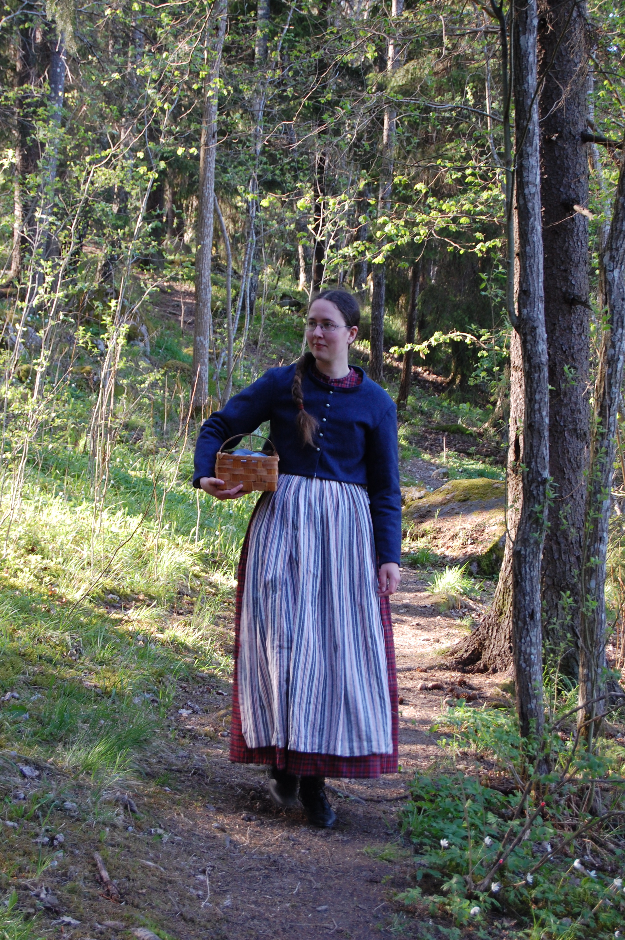 I skogen med rutig klänning, förkläde, vadmalsjacka och korg.